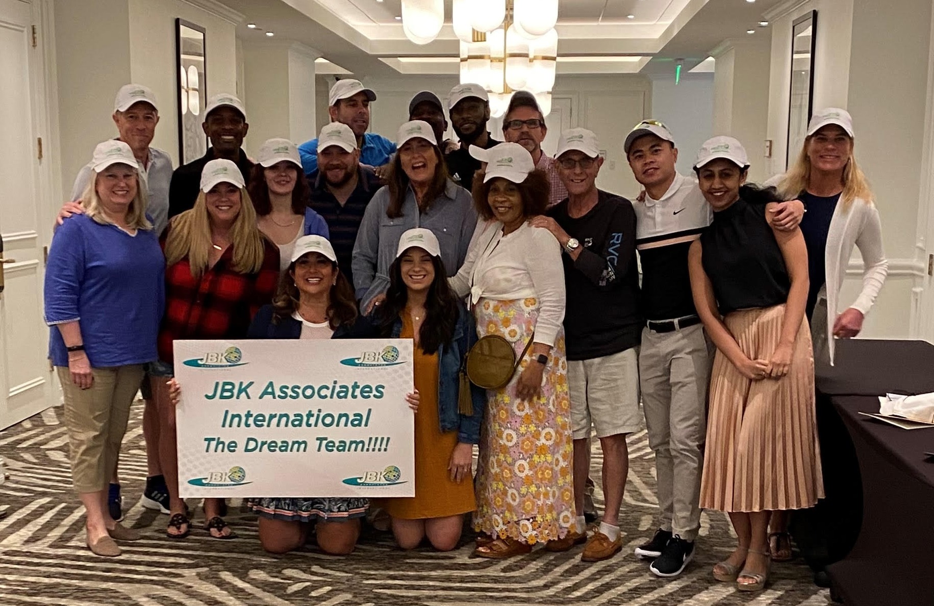 about hero JBK Dream Team (1) The JBK Associates International team wearing matching white baseball caps and holding a sign that reads "JBK Associates International The Deam Team!"
